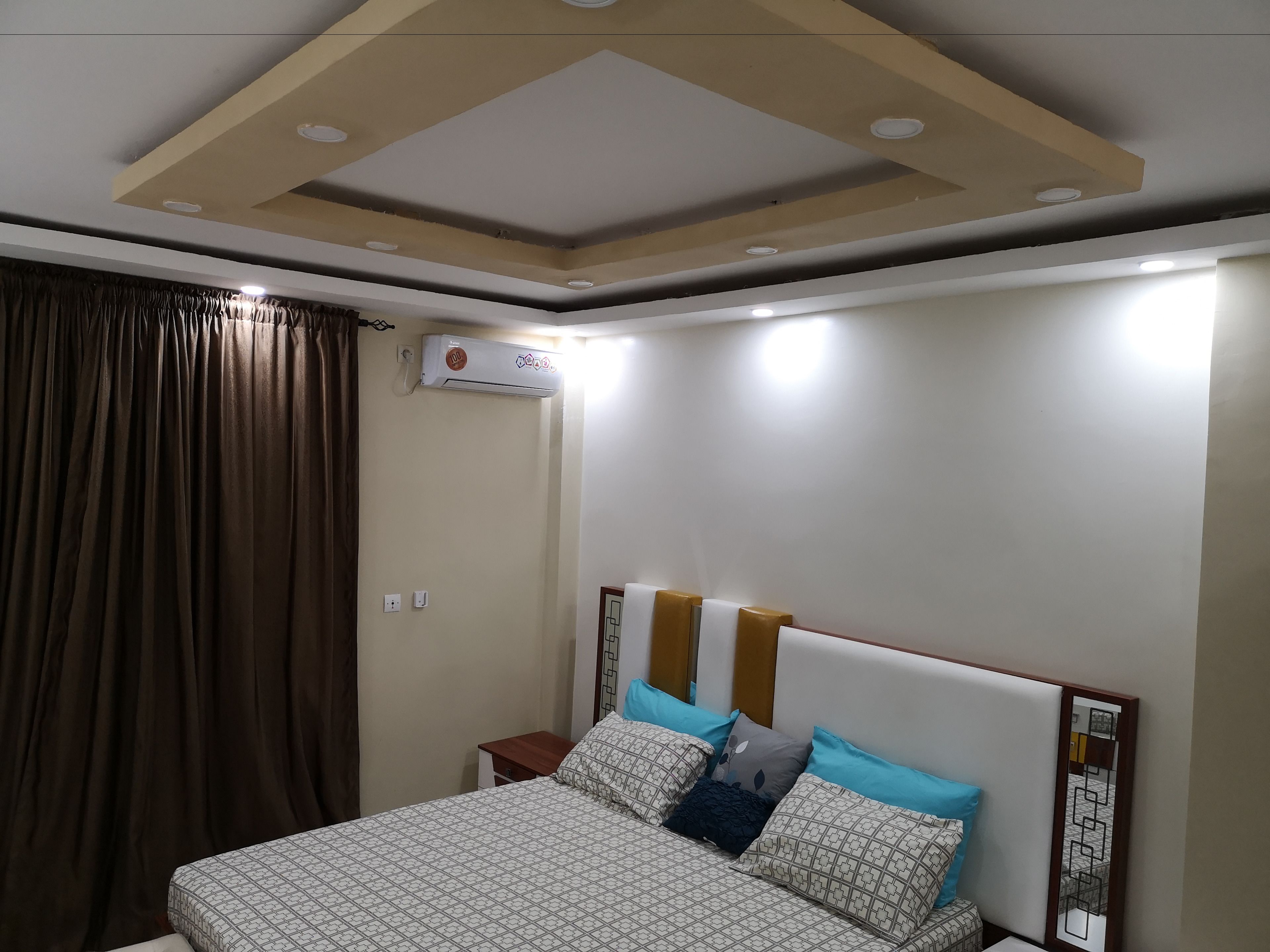 Un dormitorio con cama, cortinas, unidad de aire acondicionado e iluminación empotrada en el techo.