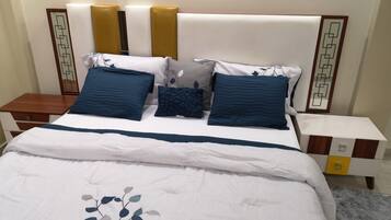Una cama bien hecha con una colcha blanca y azul, varios almohadones y una mesita de noche de madera con un cajón.