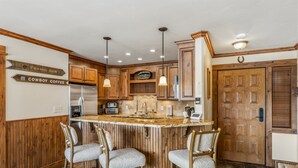 A kitchen with wooden cabinets, a central island with bar stools, a refrigerator, and a sign indicating a 'Frying Pan' and 'Cowboy Coffee.' - Bear Claw 413 Premium On Mountain Condo With Ski-In/Ski-Out Access! (Steamboat Springs)