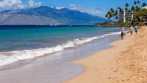 A beach with a mountain in the background, clear skies, and a few people walking along the shore.