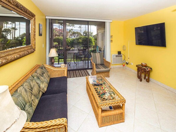A living room with a sofa, a wicker coffee table, a chair, and a television on the wall. - Suite Living w/Island Touches! Updated Kitchen, Lanai, WiFi–Kamaole Sands 1206 (KIHEI)