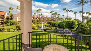 A balcony with a round table and two chairs, overlooking a well-maintained lawn with palm trees and a distant building complex.