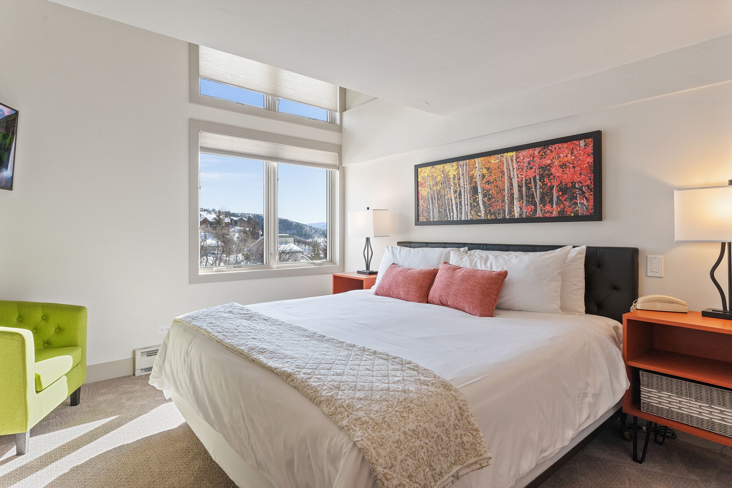 A modern bedroom with a large bed, a green chair, a painting of autumn trees, and a view of the outdoors.