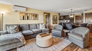 Ein modernes Wohnzimmer mit grauem Sofa, gemusterten Sesseln, einem runden Hocker und einem Essbereich mit Tisch und Stühlen.