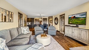 Ein geräumiges Wohnzimmer mit einem grauen Sofa, gemusterten Sesseln, einem runden Hocker und einem Holztisch. An den Wänden befinden sich ein großes Gemälde und eine Kommode.
