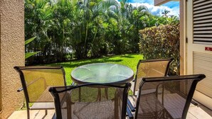 Un patio con una mesa redonda y dos sillas, con vistas a un jardín exuberante con diversas plantas y un cielo azul despejado.