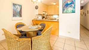 Un comedor con sillas de mimbre y una mesa redonda, una cocina al fondo y un cuadro en la pared.