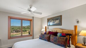 Une chambre à coucher comprenant un lit, un ventilateur de plafond, une fenêtre avec des stores, un tableau et une table de chevet avec une lampe.