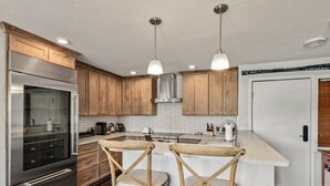 A modern kitchen with wooden cabinets, a stainless steel refrigerator, a built-in oven, and a large island with two wooden chairs. - Bear Claw 109 on Mountain Condo with Ski-In/Ski-Out Access! (Steamboat Springs)