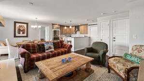 A spacious living room with a wooden coffee table, a plaid sofa, and a green armchair. A kitchen with wooden cabinets and stainless steel appliances is visible in the background. - Bear Claw 109 on Mountain Condo with Ski-In/Ski-Out Access! (Steamboat Springs)
