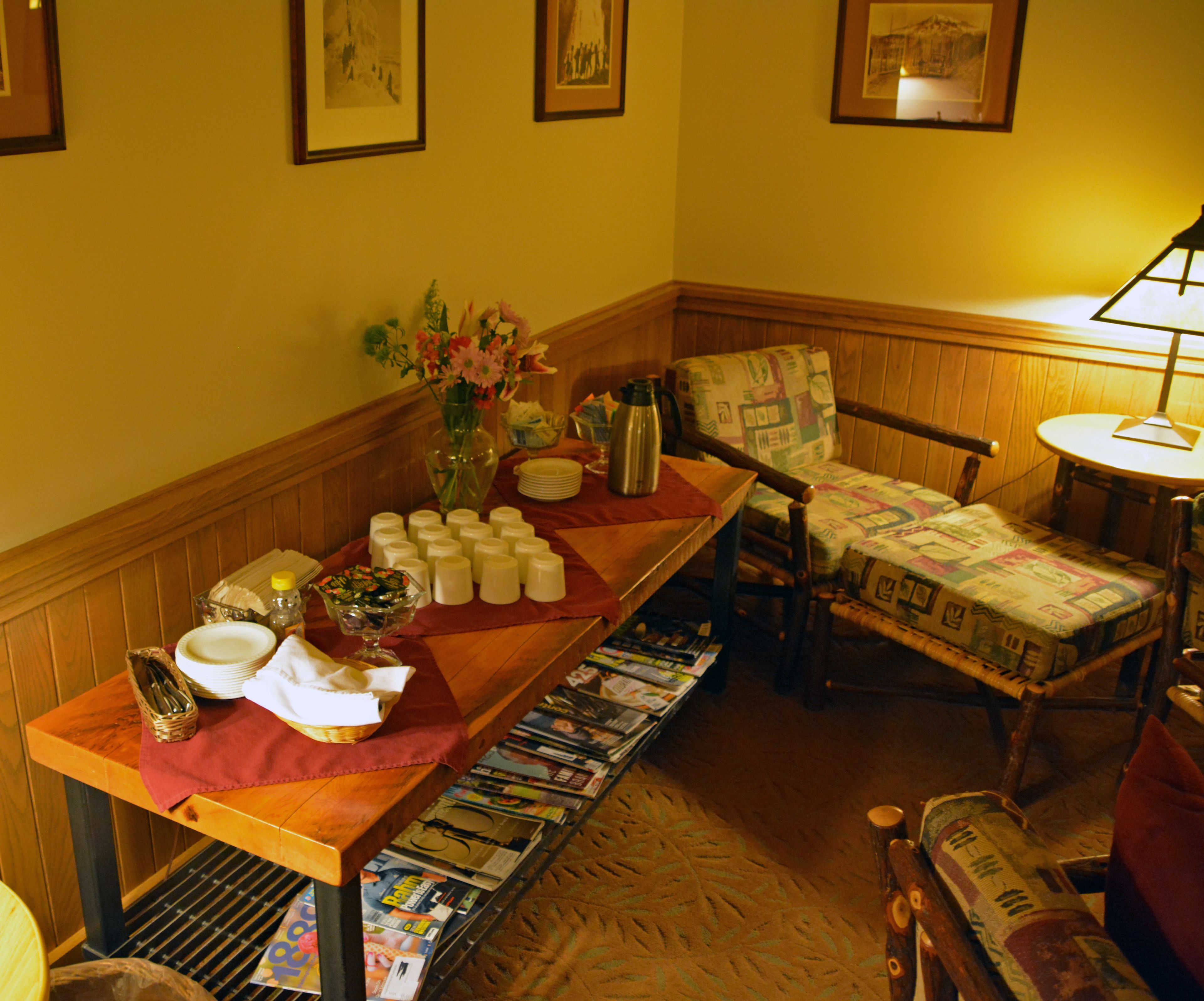 A corner of a room with a wooden table set for a meal, a chair with a quilted cover, and a magazine rack filled with magazines.