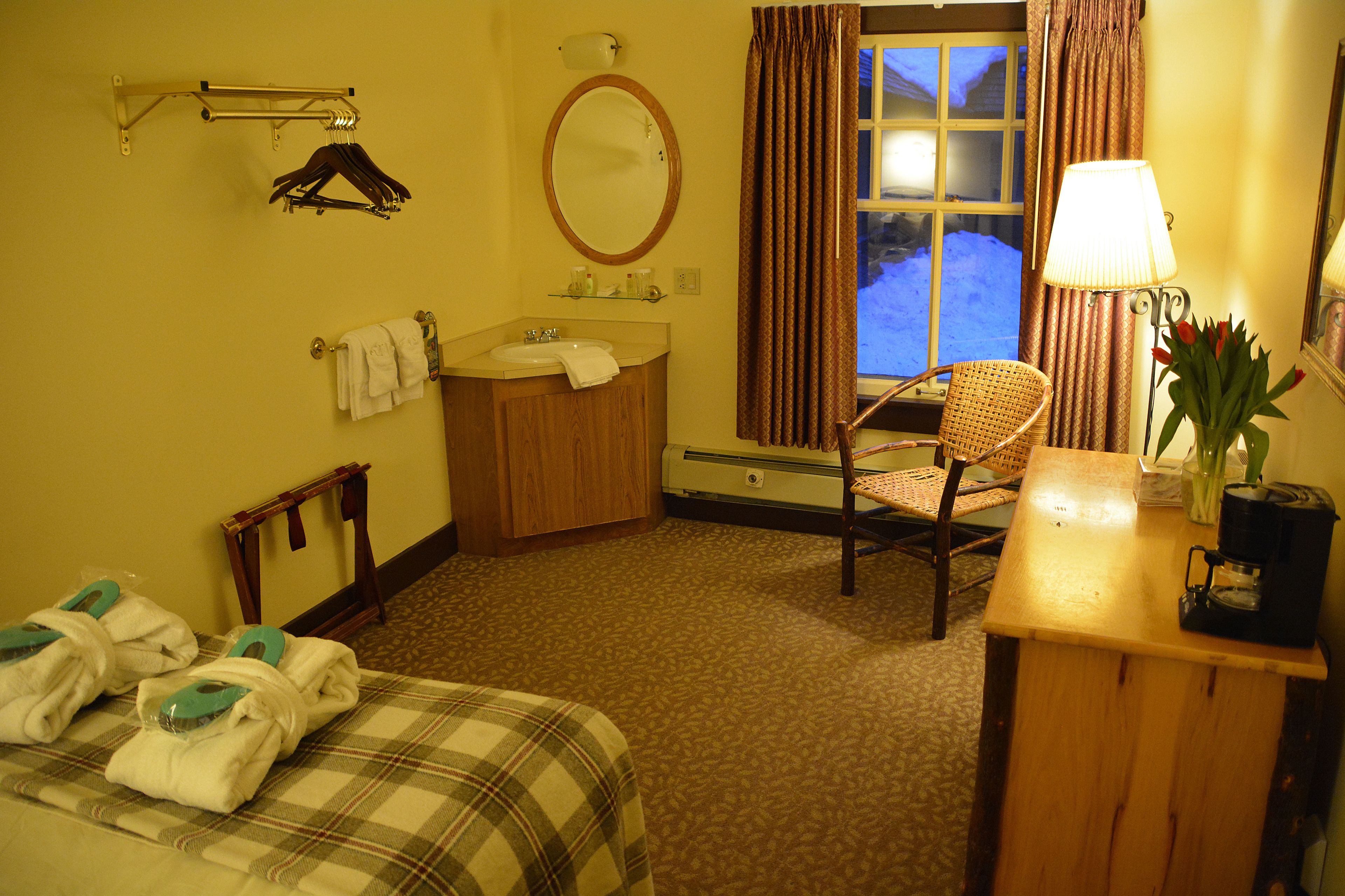 A hotel room with a bed, a chair, a desk with a lamp, a mirror, a window with curtains, and a bathroom with a sink and towel rack.