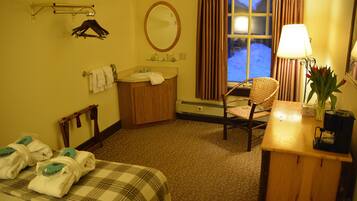 A hotel room with a bed, a chair, a desk with a lamp, a mirror, a window with curtains, and a bathroom with a sink and towel rack.