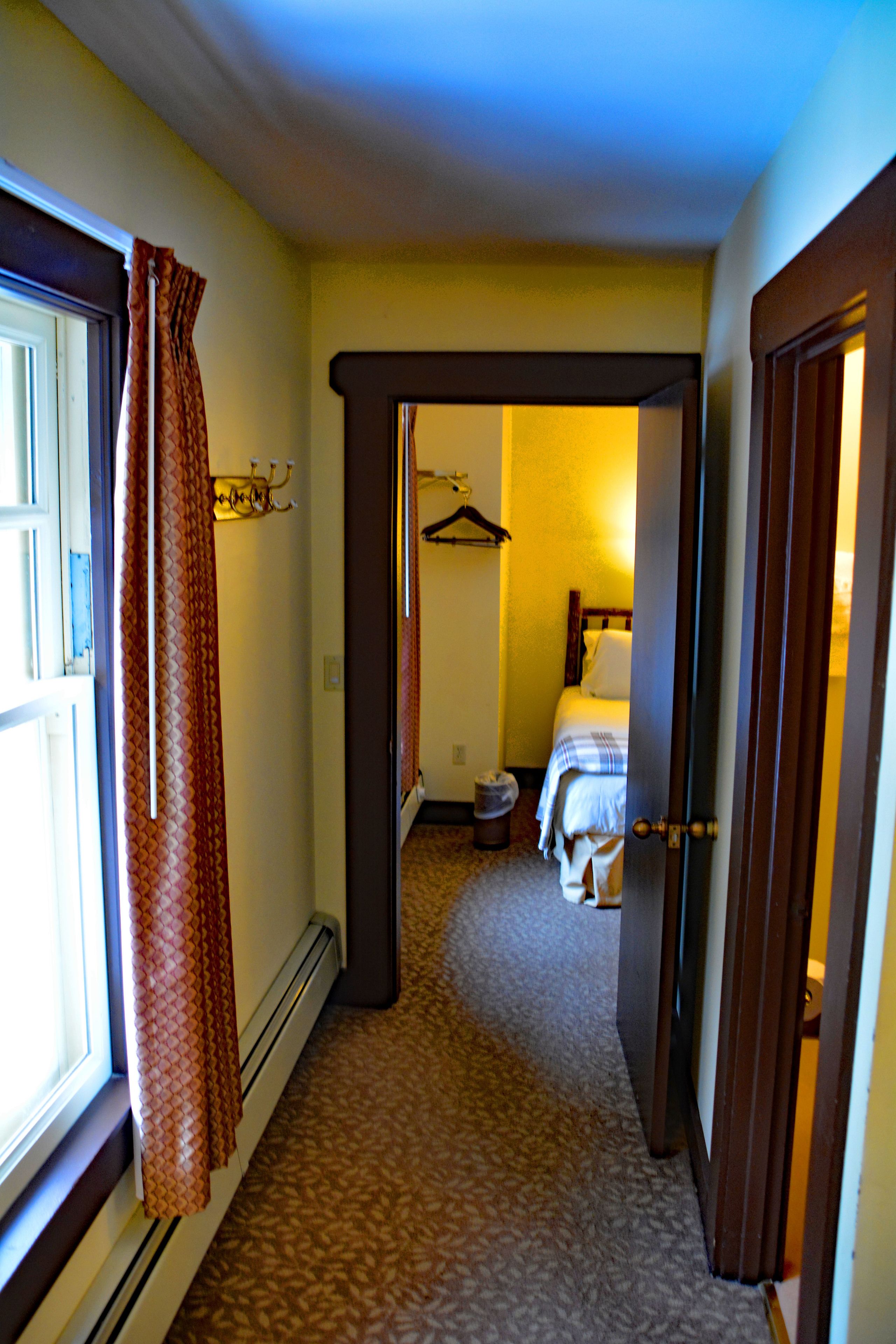 A narrow hallway with a carpeted floor leading to a bedroom with a bed and a hanging coat rack.