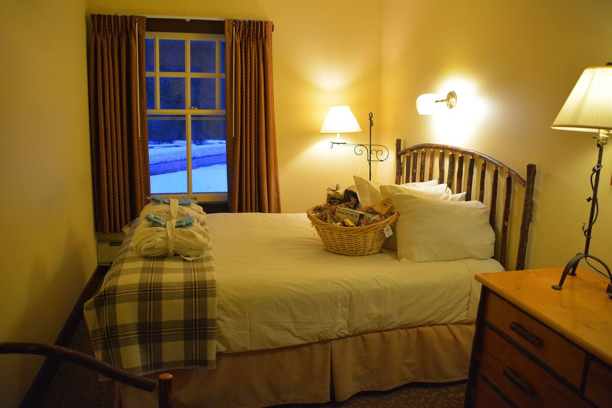 Ett hotellrum med en säng, ett sängbord, en lampa, ett fönster med gardiner och utsikt över omgivningen.