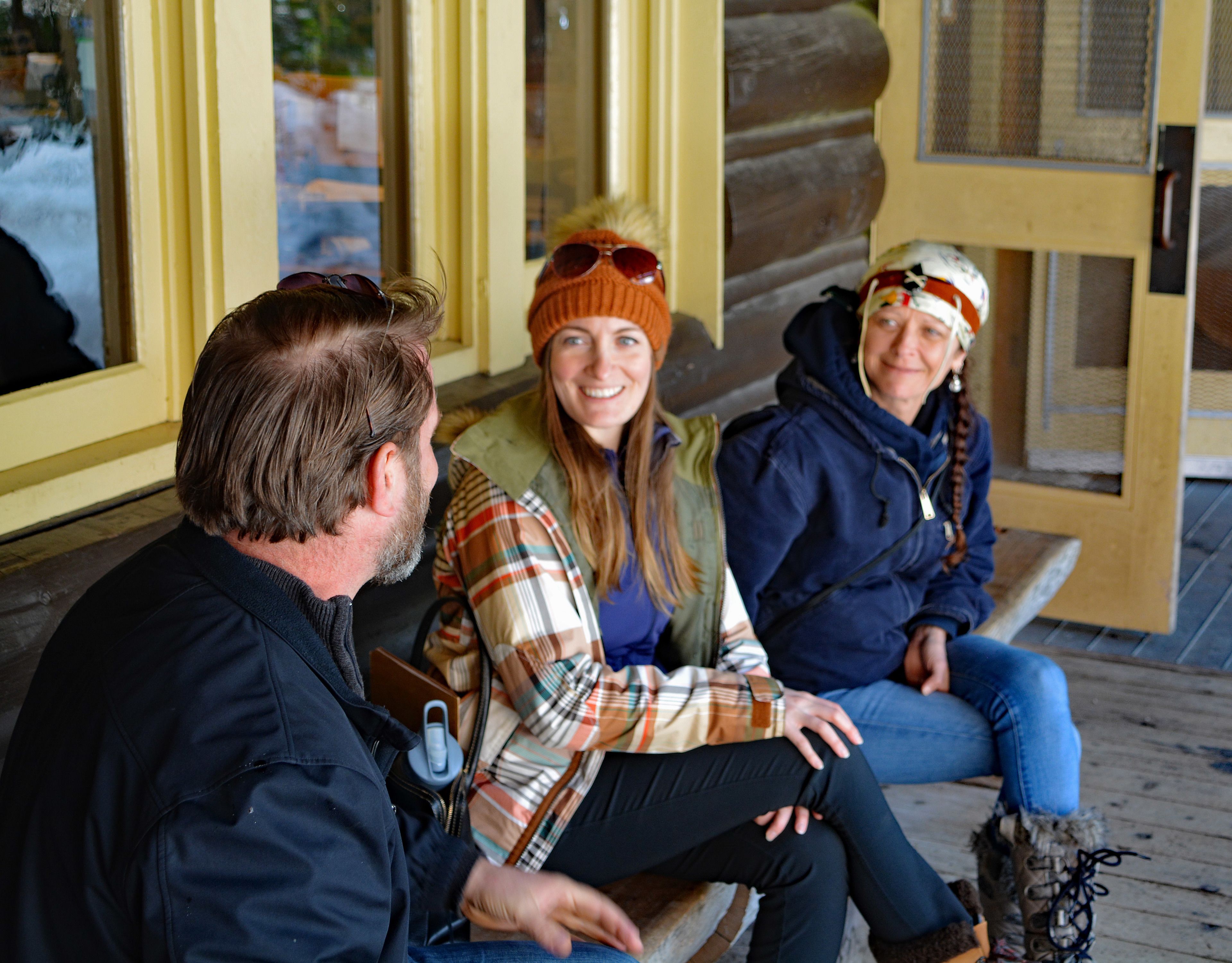 Three people sitting on a wooden bench outside a building with yellow walls and large windows.