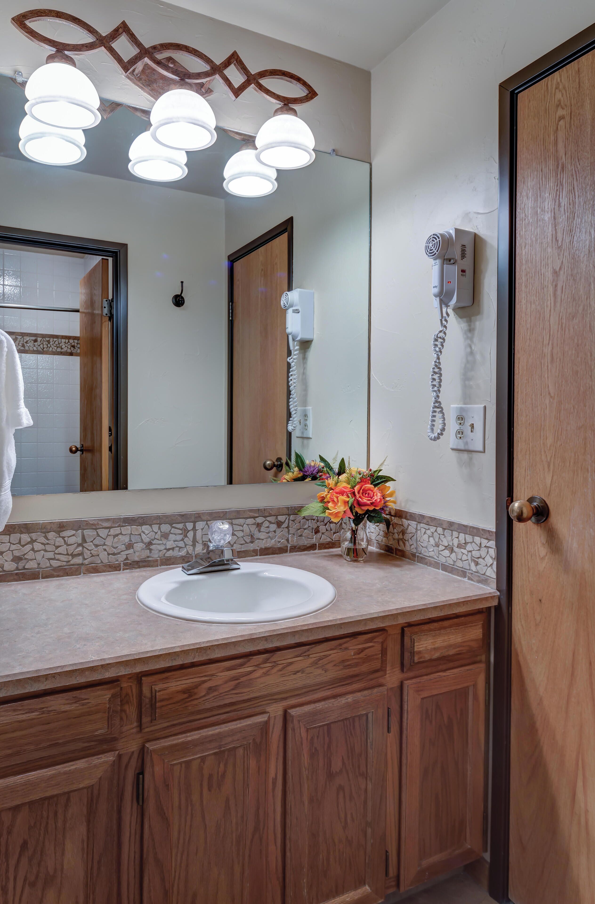 Une salle de bain avec un meuble-lavabo en bois, un lavabo blanc, un miroir et un sèche-cheveux fixé au mur.
