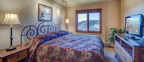A bedroom with a bed, a nightstand, a lamp, a dresser, a television, a window with a view, and a framed picture on the wall.