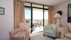 A room with a patterned armchair, a blue patterned chair, and a wicker sofa. There is a balcony with a table and chairs, and a statue on a side table.