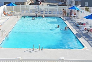 A rectangular swimming pool with multiple lanes, surrounded by white fences and shaded by blue umbrellas.