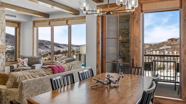 A dining area with a wooden table and chairs, a chandelier, and large windows offering a mountain view.