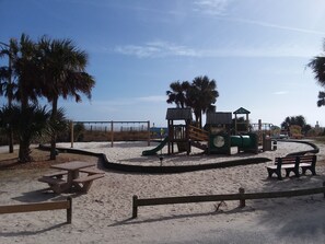 A sandy playground with a slide and climbing structures, surrounded by palm trees and picnic tables.