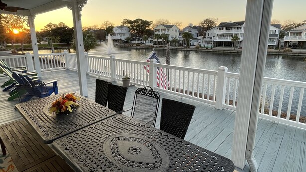 Une terrasse couverte, aménagée avec du mobilier d’extérieur, offrant une vue sur un lac et sur des maisons situées au loin.