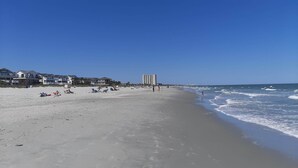 Une vaste plage de sable où se trouvent quelques personnes et des chaises de plage, un grand immeuble en arrière-plan et des vagues qui se dirigent vers le rivage.