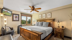Une chambre avec un lit en bois, un ventilateur de plafond, une grande fenêtre et un téléviseur fixé au mur.