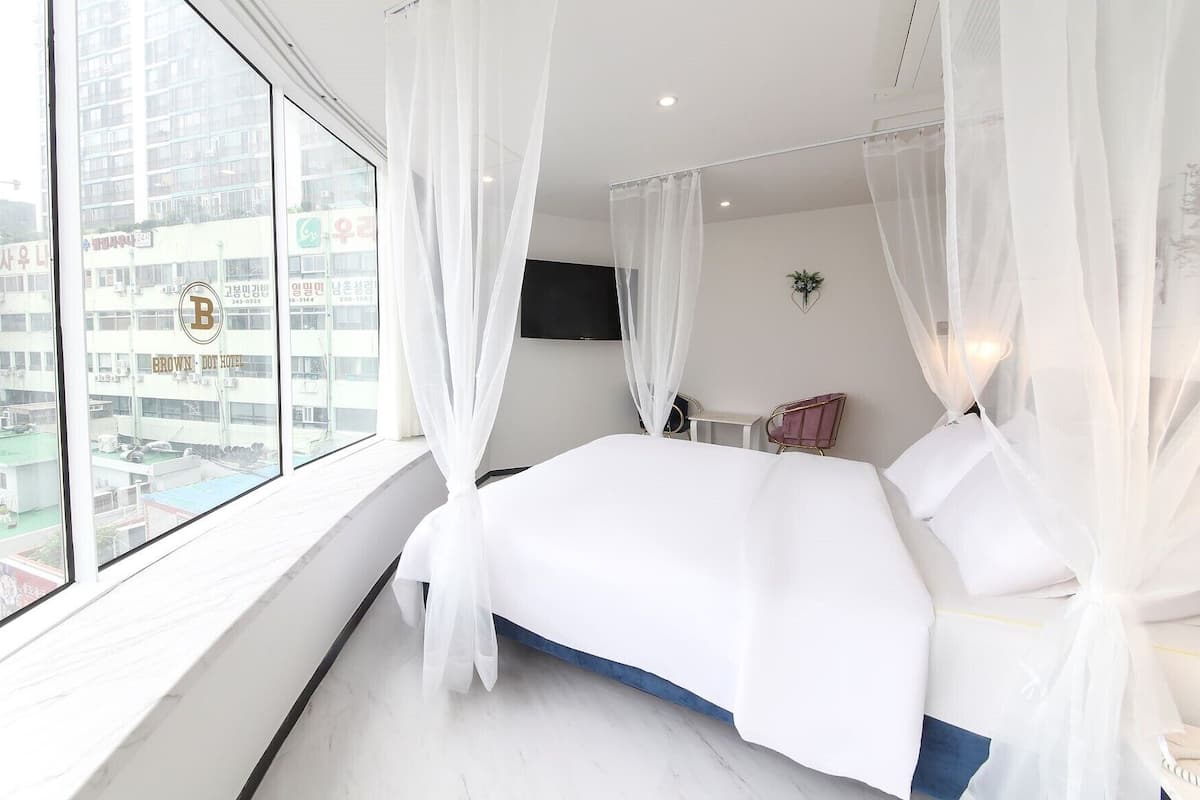 Une chambre d’hôtel avec un grand lit, des rideaux blancs, un téléviseur à écran plat et une vue sur le paysage urbain.