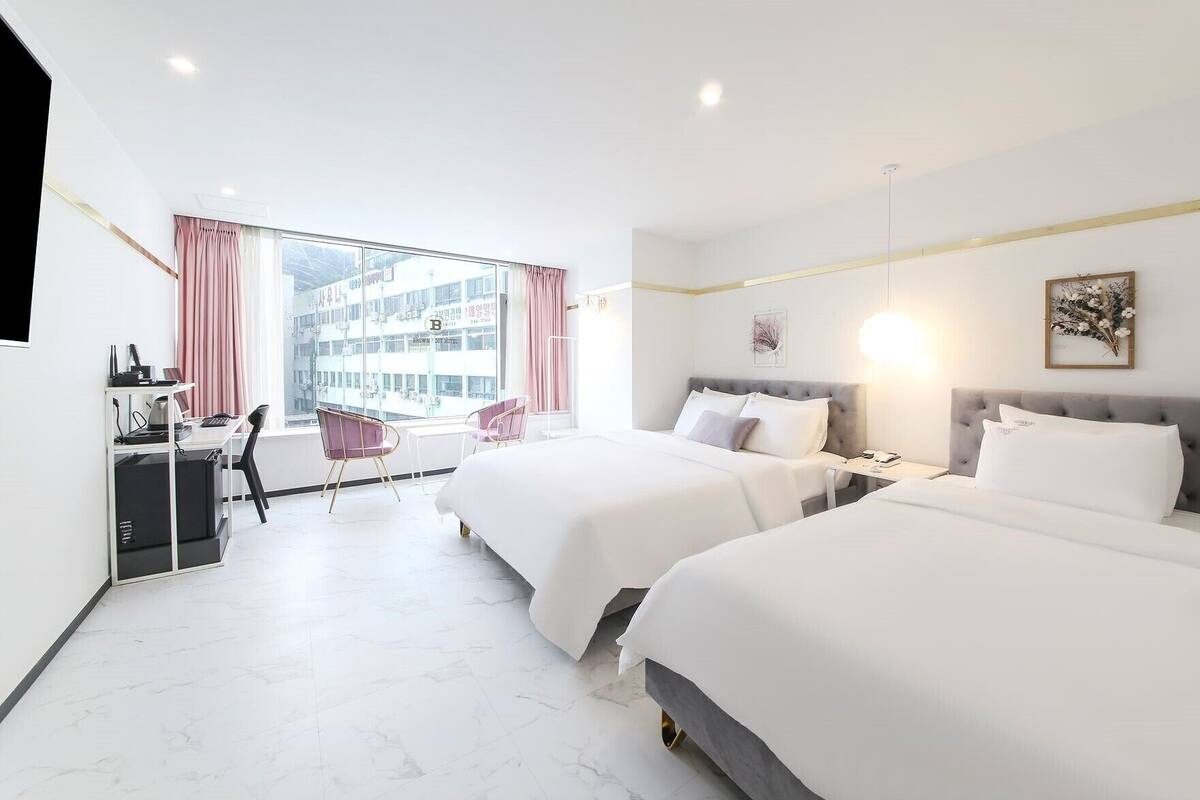 Une chambre d’hôtel avec deux lits, un bureau équipé d’un ordinateur et une vue sur un immeuble par de grandes fenêtres.