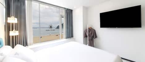 Hotelkamer met een groot bed, een aan de muur bevestigde televisie en uitzicht op het strand door een groot raam.
