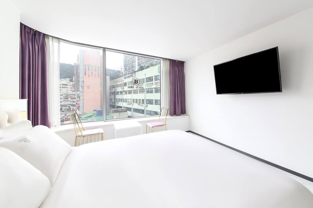 Une chambre d’hôtel avec un grand lit, un téléviseur fixé au mur, une fenêtre avec des rideaux et une vue sur le paysage urbain.