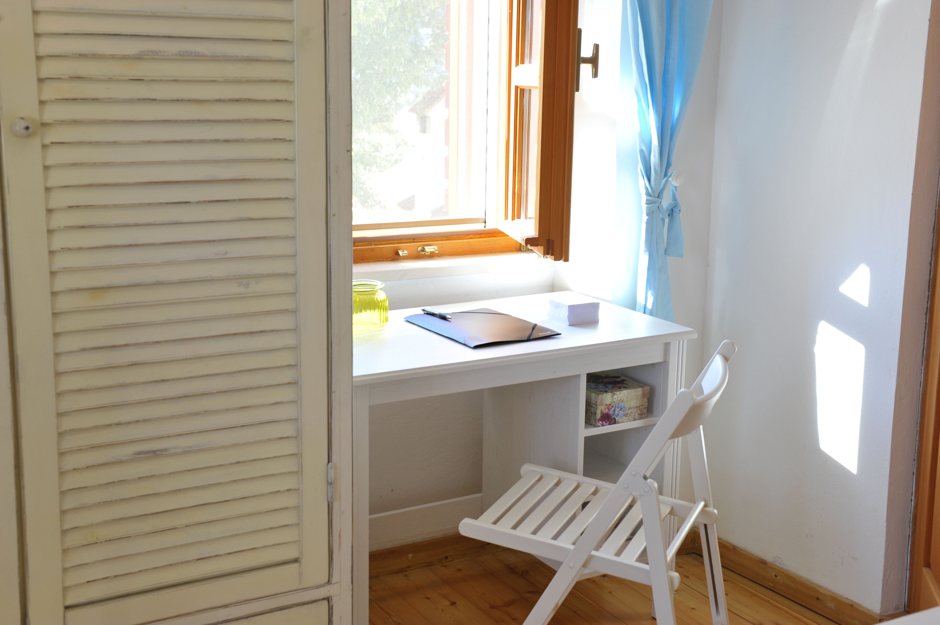 A small, white desk with a chair, a window with curtains, and a shelf below.