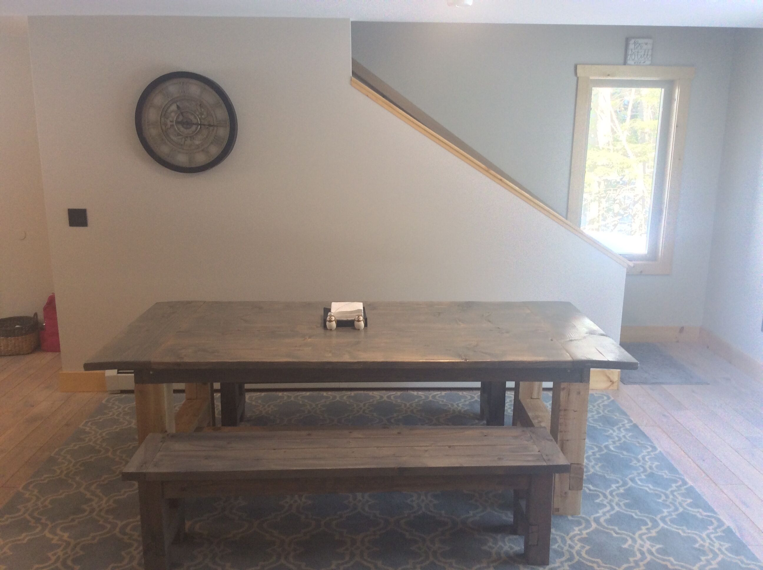 A spacious room with a large wooden table and bench, a clock on the wall, and a window with a view of trees.