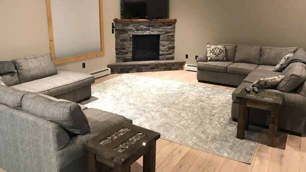 A living room with a stone fireplace, two grey sofas, a small wooden coffee table, and a large window with blinds.