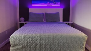 Ein Schlafzimmer mit einem Bett, zwei Kissen, einem Nachttisch, einem an der Wand montierten Fernseher und einem Bild von einer Strandansicht.