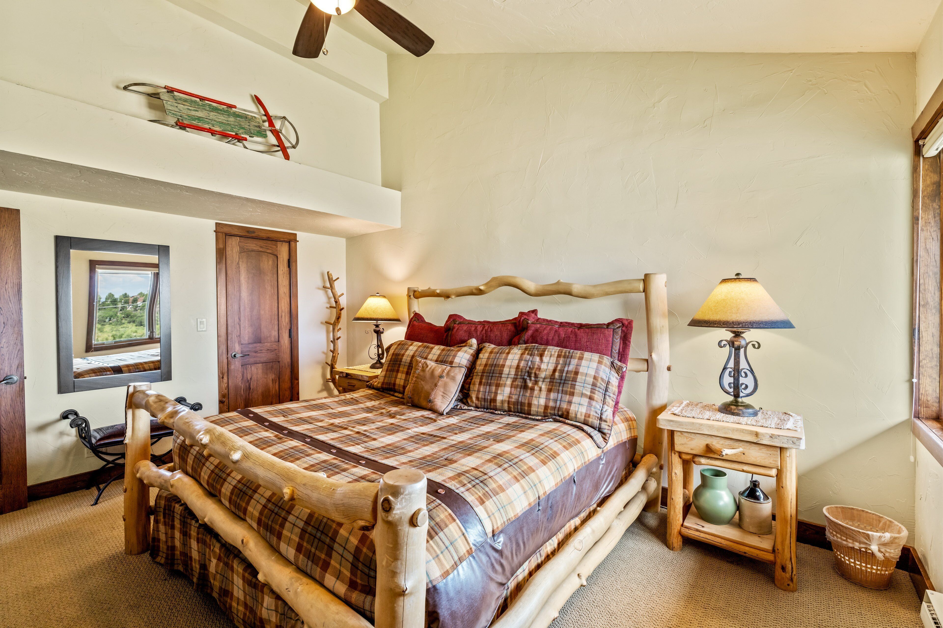 A bedroom with a wooden bed frame, plaid bedding, two bedside lamps, and a small wooden nightstand with a lamp and a vase.