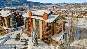 A multi-story hotel with a snow-covered roof, surrounded by a parking area and a mountainous backdrop. - Bear Claw 404 Premium On Mountain Condo With Ski-In/Ski-Out Access! (Steamboat Springs)