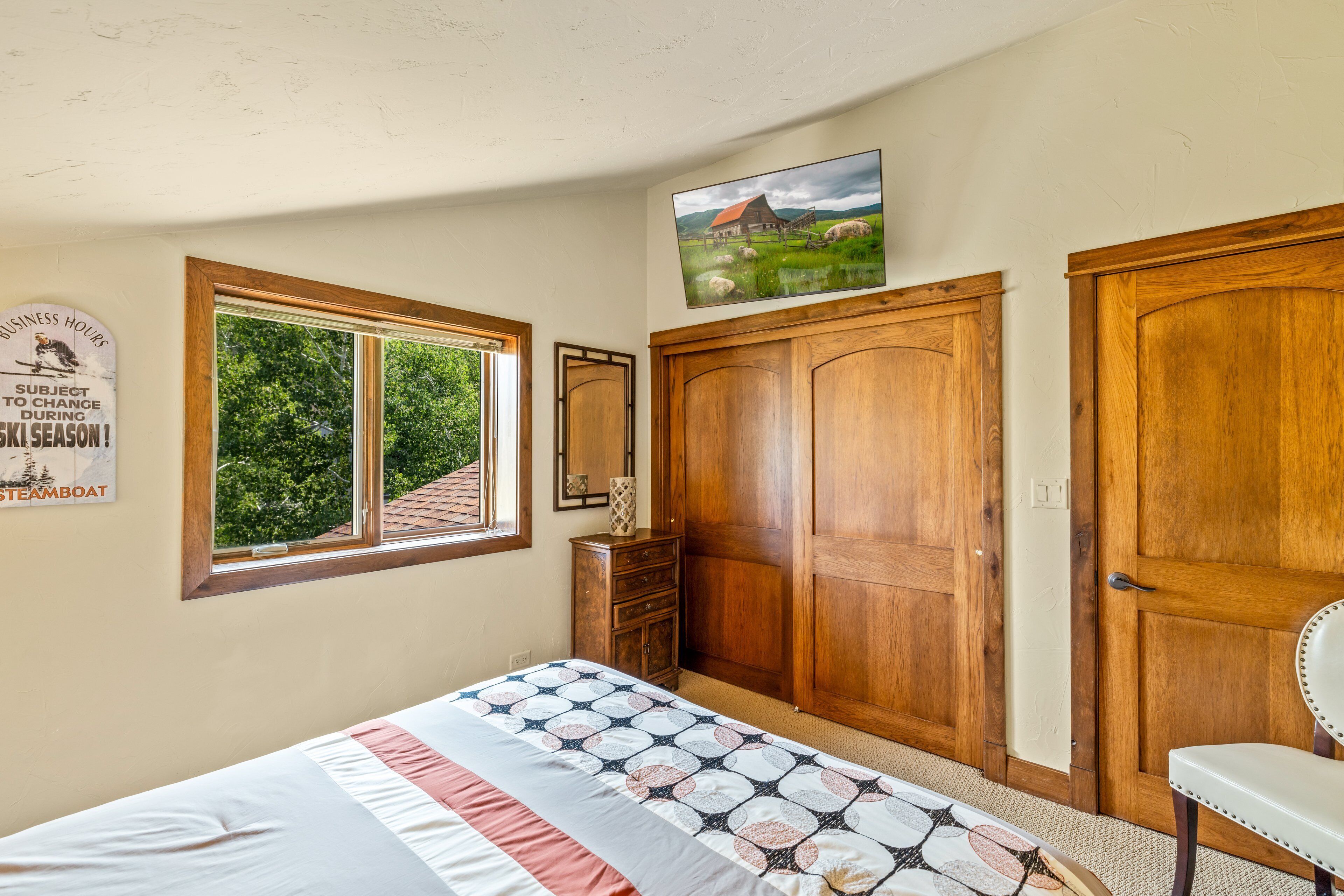 A bedroom with a bed, a wooden wardrobe, a mirror, a window with a view of greenery, and a wall-mounted picture.
