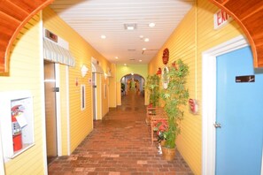 Un pasillo de hotel con paredes amarillas, piso de ladrillo y una puerta azul. Se observan carteles que indican números de habitaciones y una salida.