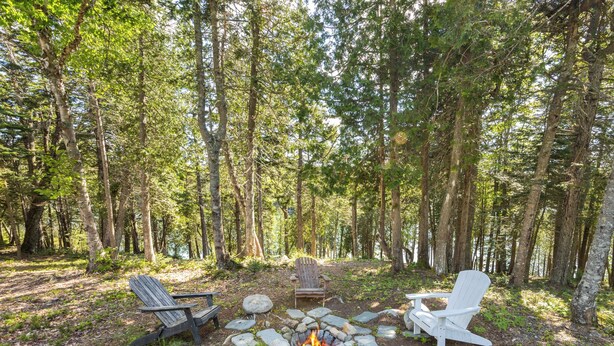 A forested campsite with a stone fire pit, two Adirondack chairs, and a white chair.