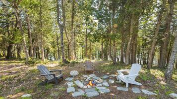 Un terrain de camping boisé, doté d’un foyer en pierre, de deux fauteuils Adirondack et d’une chaise blanche.