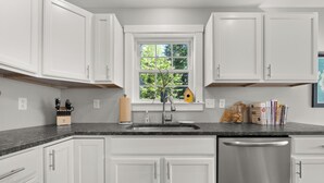 A kitchen with white cabinets, a granite countertop, a stainless steel dishwasher, a window with a view of greenery, and a birdhouse on the windowsill.