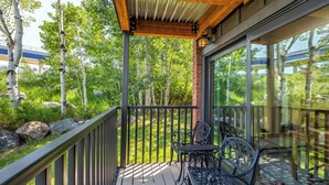Une terrasse en bois avec des rampes en métal, deux chaises en fer forgé et une porte vitrée donnant sur un jardin luxuriant.