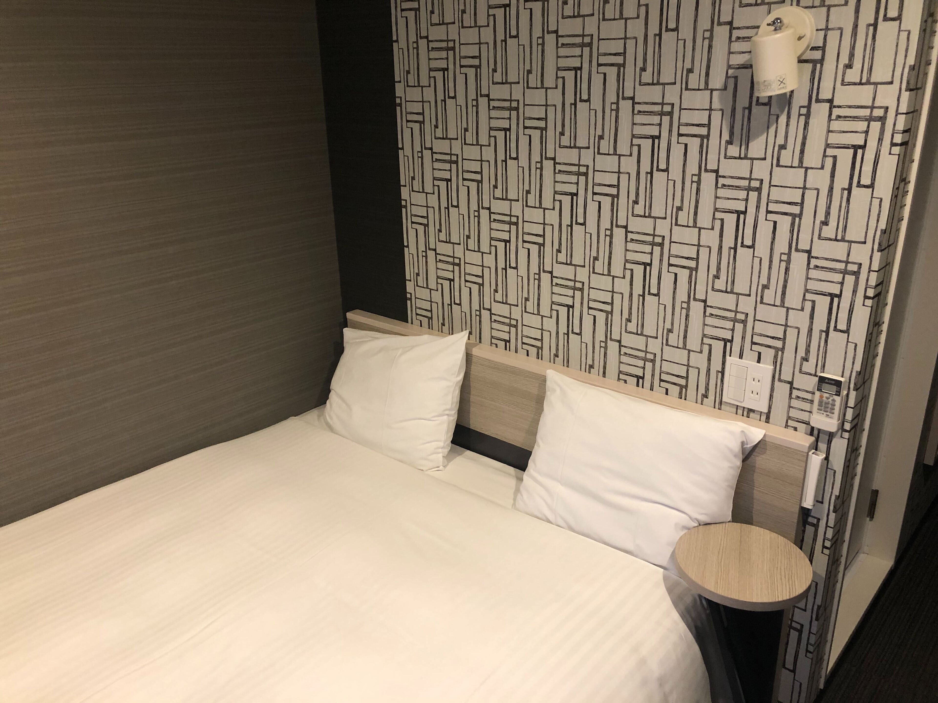 A hotel room with a bed, two pillows, a bedside table, and a wall with a geometric pattern.