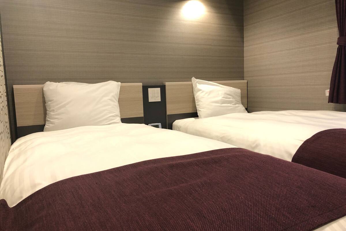 Habitación de hotel con dos camas, cada una con ropa de cama blanca y cabecero. Hay una lámpara de pared sobre las camas y una cortina oscura a la derecha.