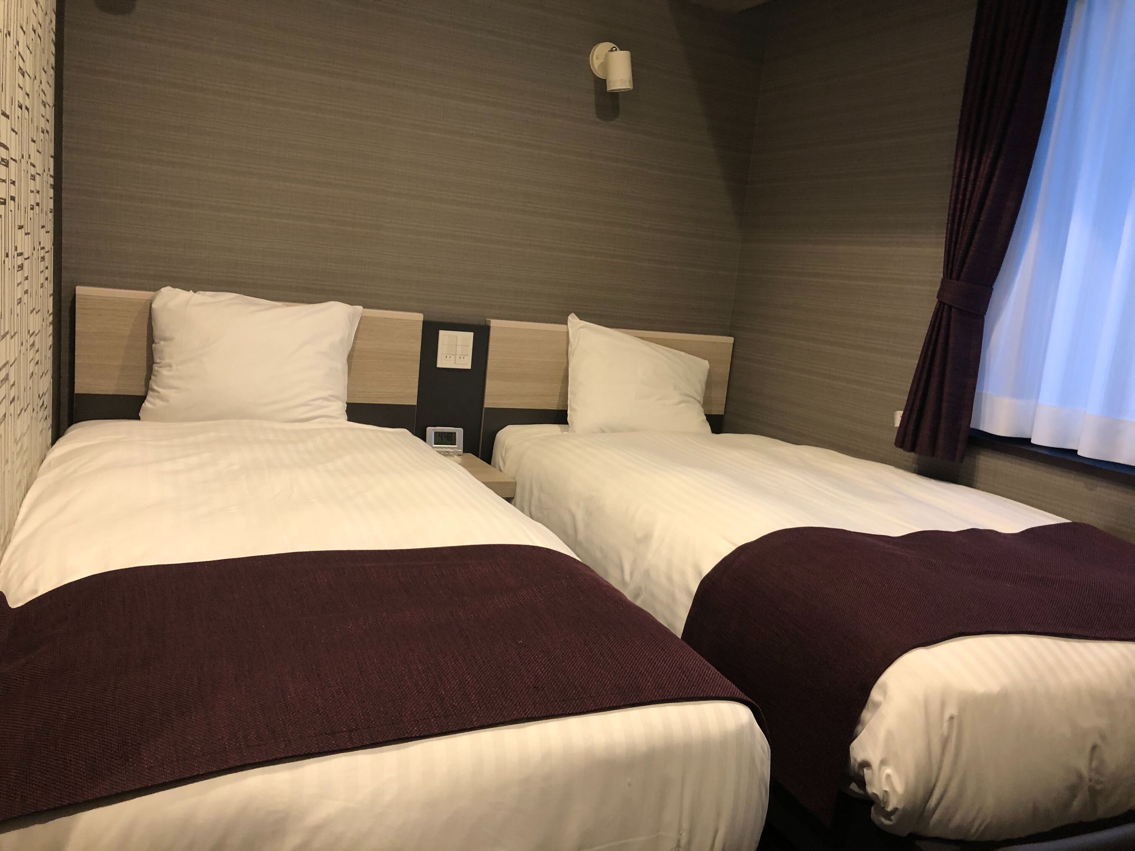 Habitación de hotel con dos camas, cada una con ropa blanca y una manta marrón oscuro. Hay una lámpara de pared sobre las camas y una ventana con cortinas.
