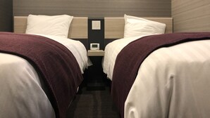 A hotel room with two beds, each with white linens and a brown blanket. There is a nightstand between the beds with a small white device on it. The room has a modern design with a wooden headboard and a light fixture above the beds.