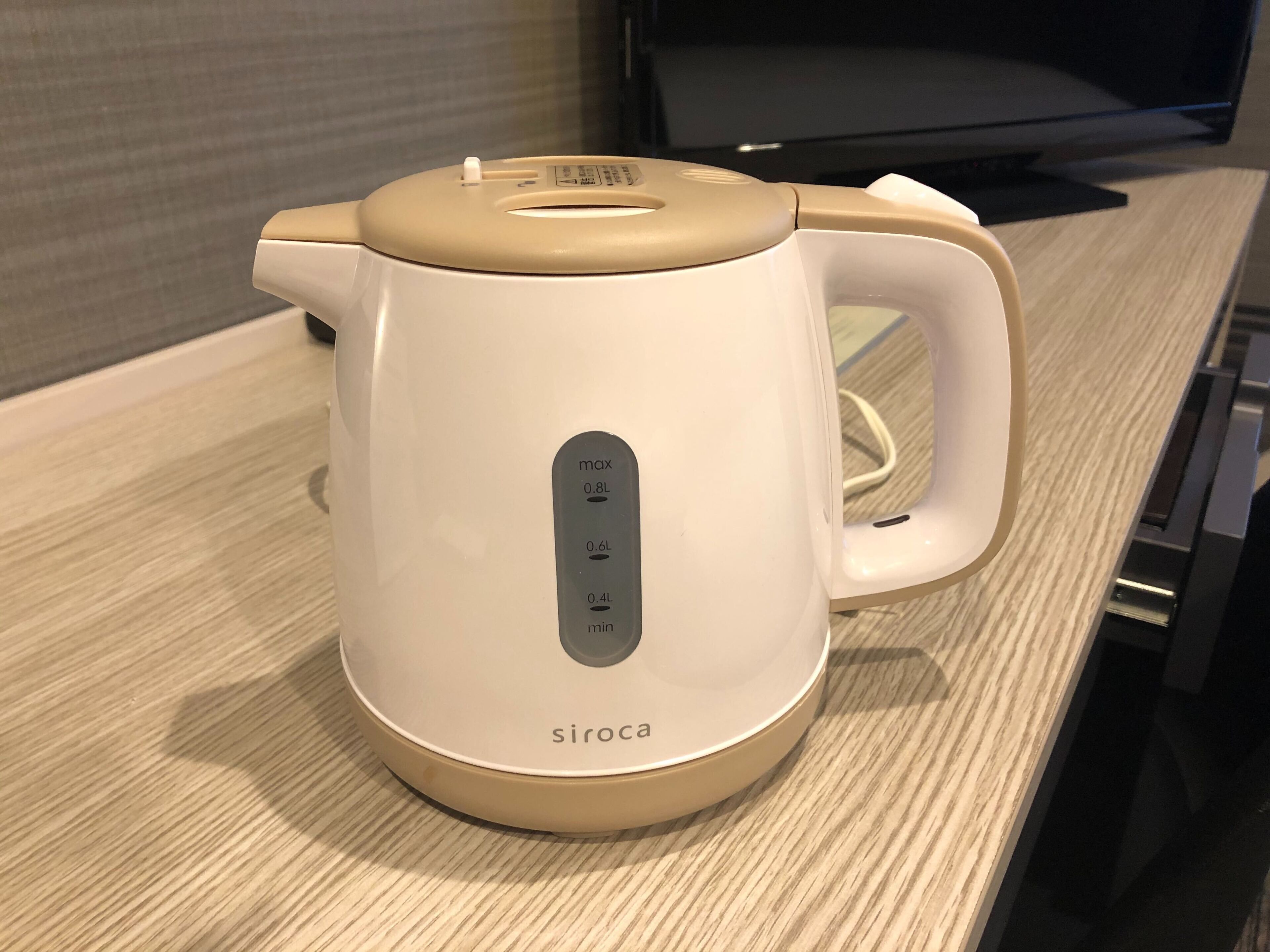 Uma chaleira elétrica branca e bege com visor digital e cabo, apoiada em uma superfície de madeira.